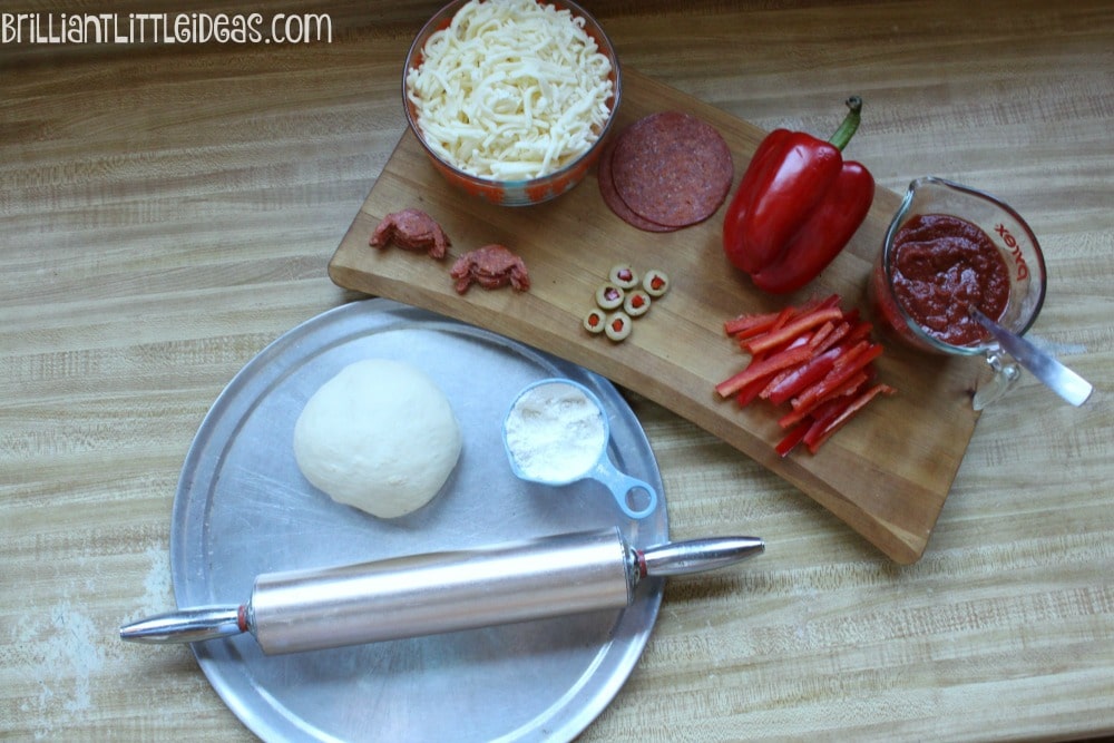On a tan kitchen counter top sits a ball of pizza dough, cup of flour and rolling pin sitting on a pizza pan. Next to the pan is a wooden cutting board with pepperonis, cheese, bell peppers, and pizza sauce. 
