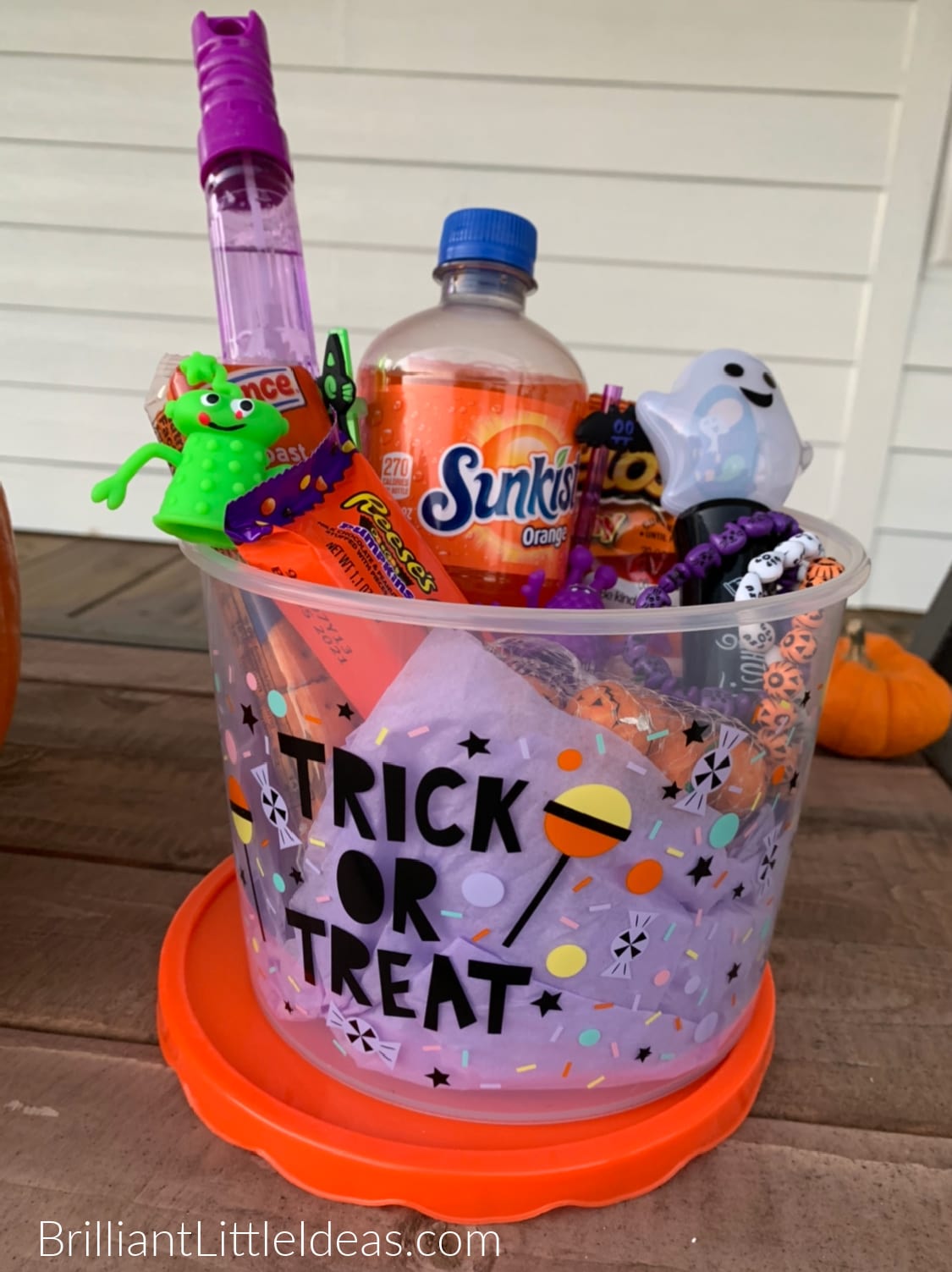 A filled clear Trick or Treat bucket sits ontop of its orange lid. The bucket is filled with orange candy, drinks, and toys. All on a dark wood background. 