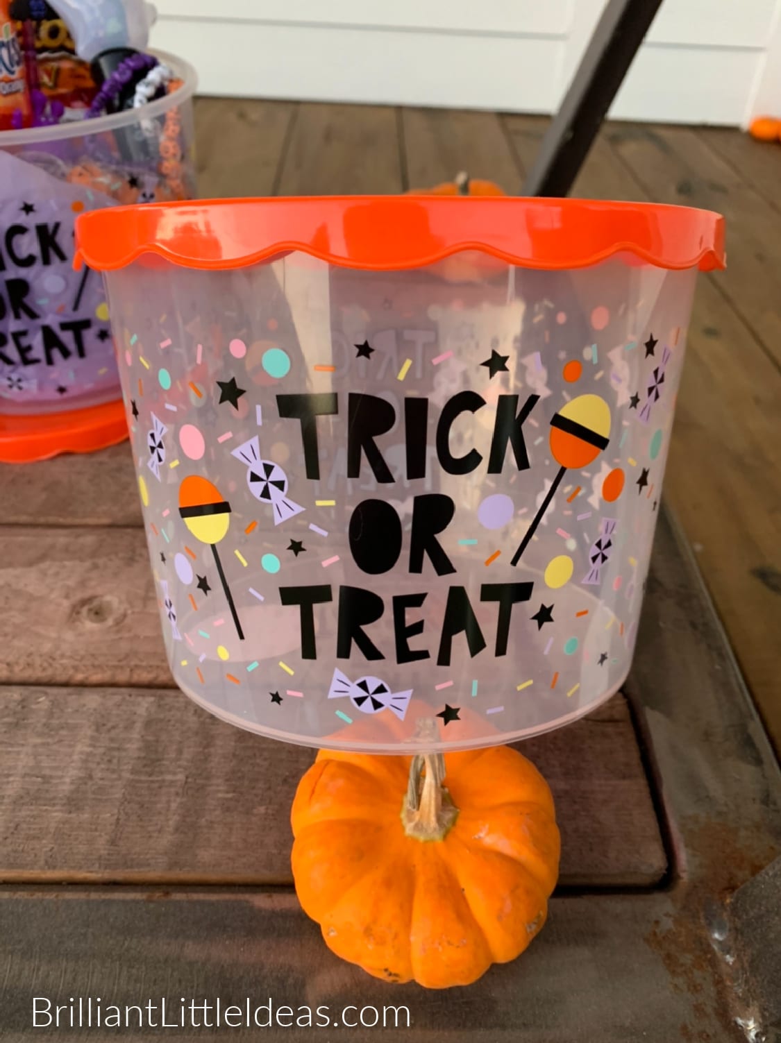 A clear bucket with a lid that says Trick or Treat and colorful candy throughout the bucket. The bucket sits next to a small pumpkin. 